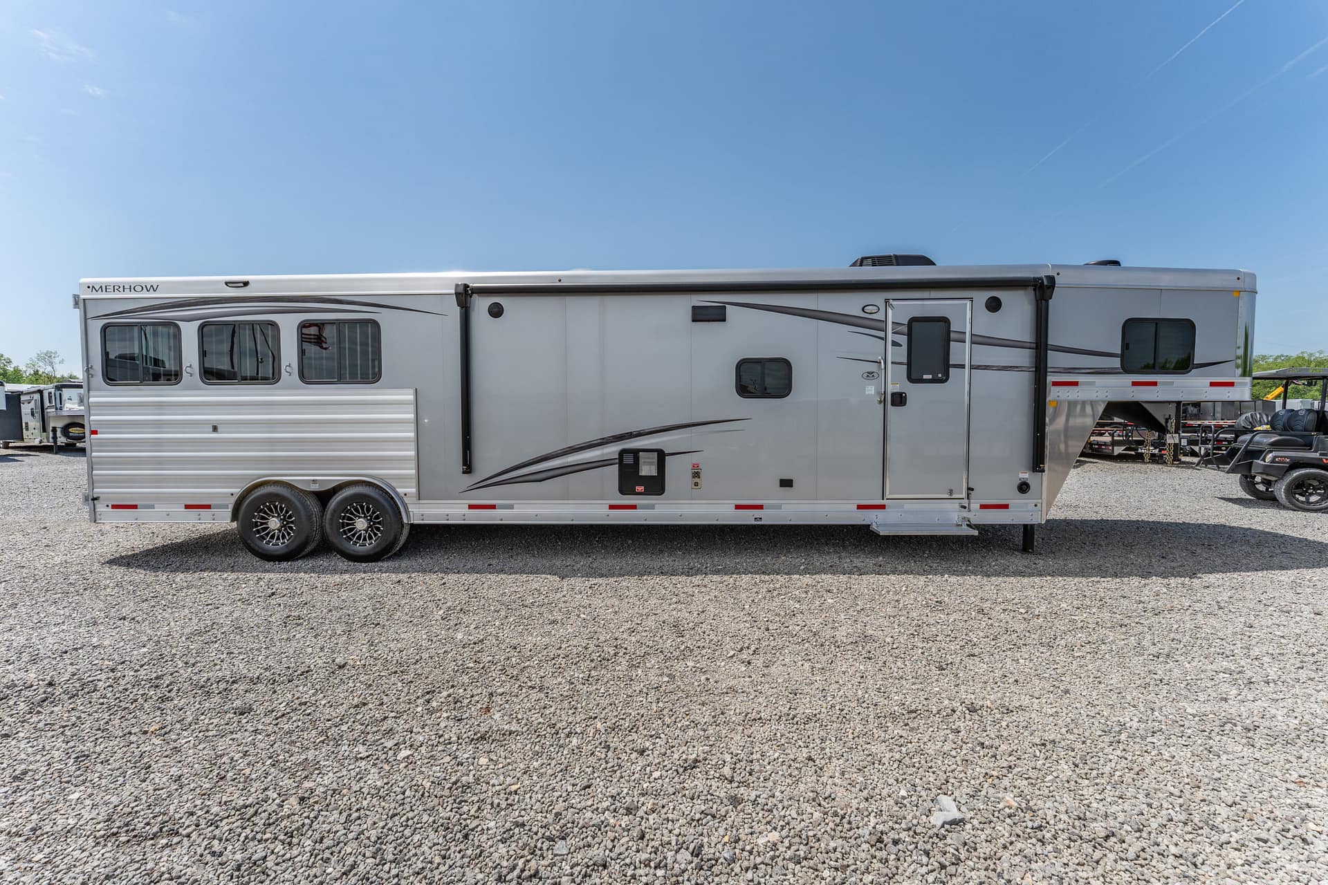 Silver Merhow fifth-wheel trailer parked on a gravel lot under a clear blue sky, viewed from the side.