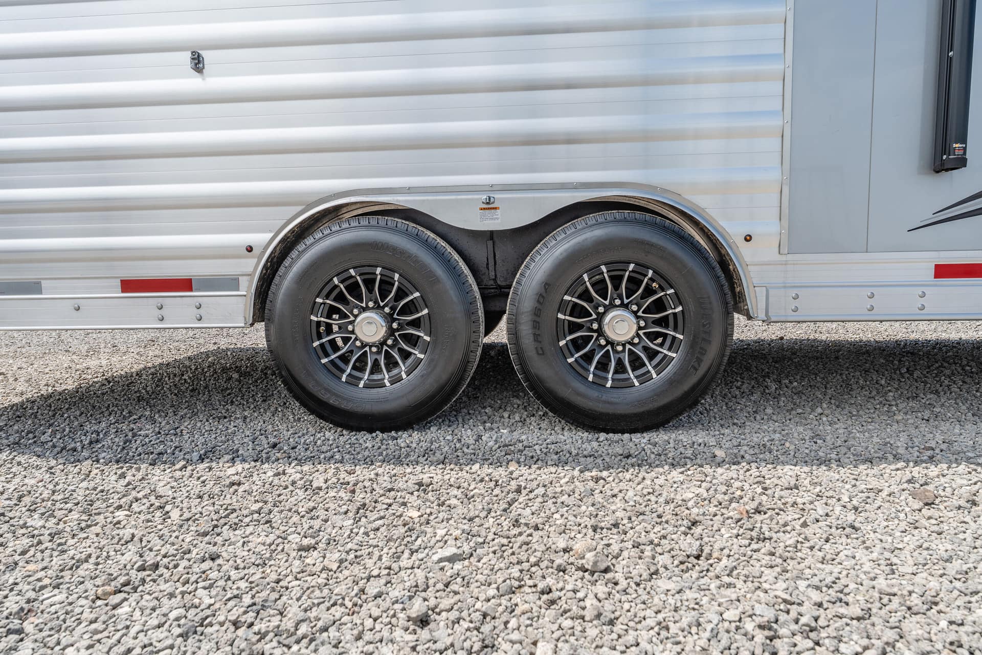Side view of a silver travel trailer with dual-axle wheels on a gravel surface.