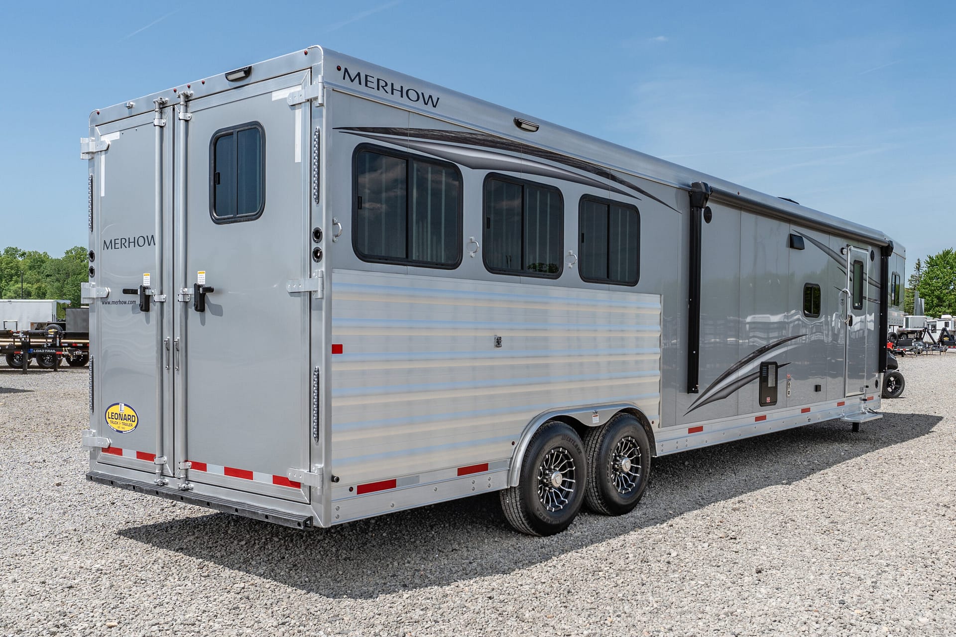 Silver Merhow horse trailer with multiple windows and dual-axle wheels parked on a gravel lot under a clear blue sky