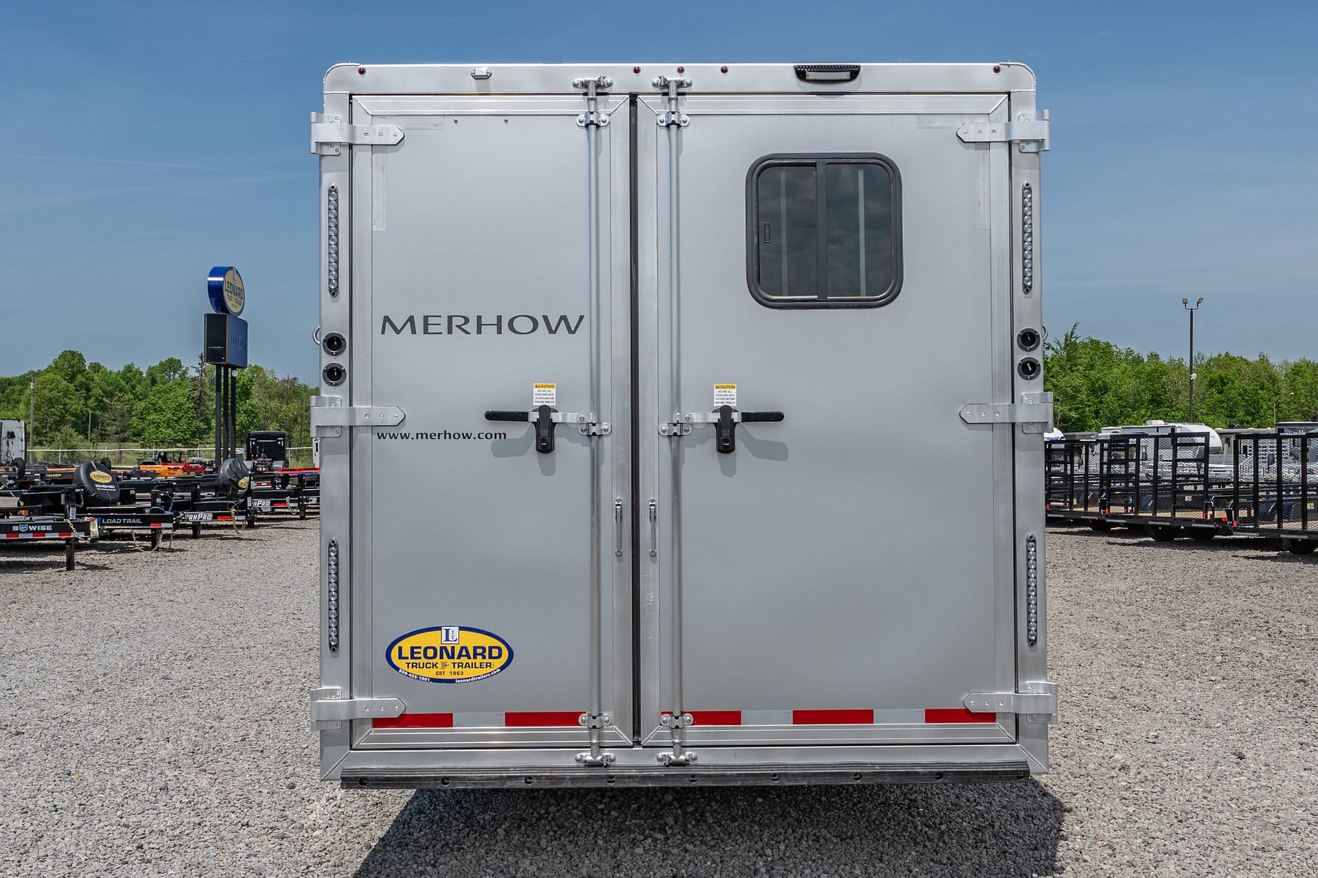 Rear view of a silver Merhow trailer doors closed, with a small window and Leonard logo on a gravel lot.