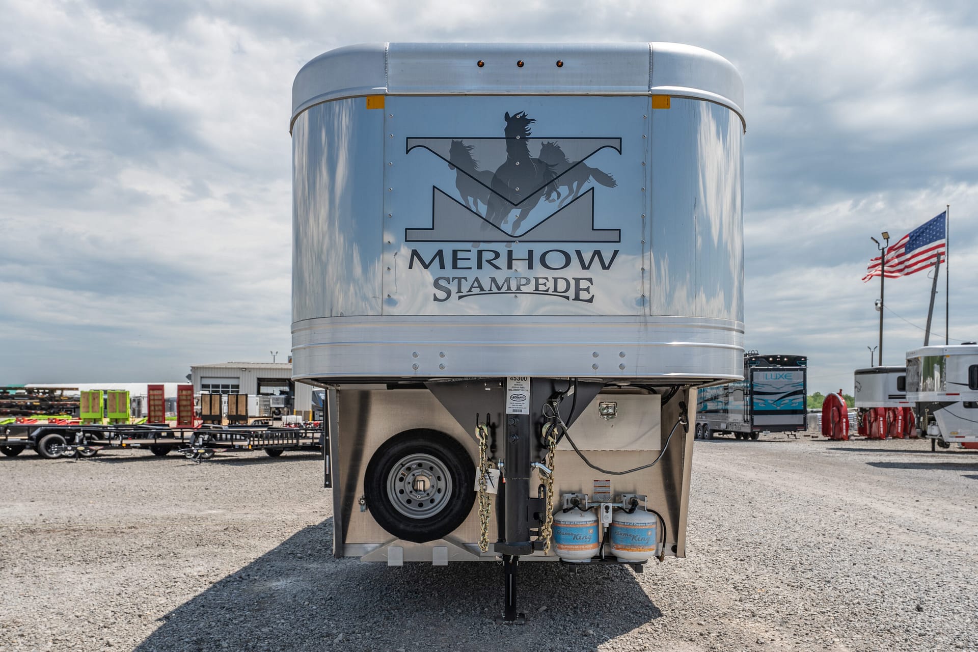 Rear of a silver Merhow Stampede horse trailer with its logo, parked in a gravel lot; American flag visible in the background.