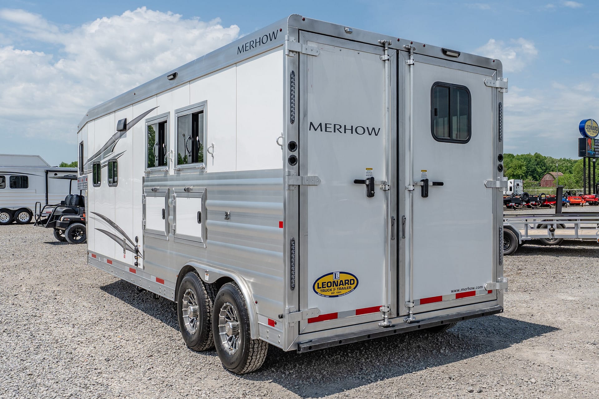 White Merhow horse trailer with rear double doors, side windows, and storage compartments on a gravel lot.