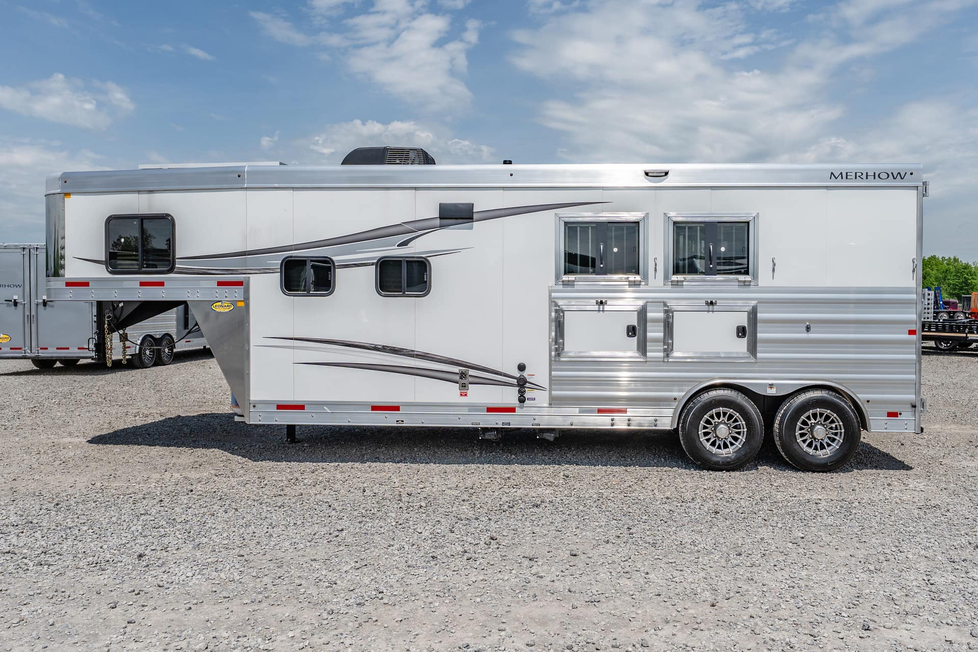 Side view of a large silver Merhow horse trailer on a gravel lot under a blue sky.