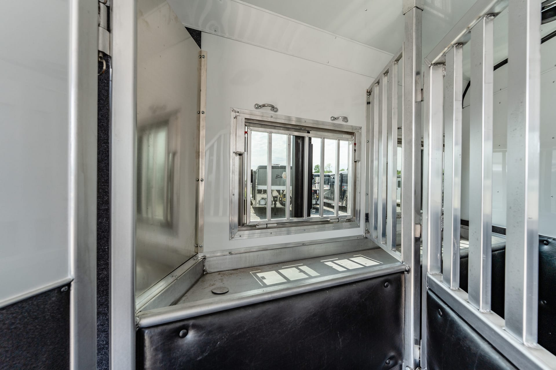 Interior of a metal trailer with vertical aluminum framing rails and a small rectangular window showing parked trucks outside.