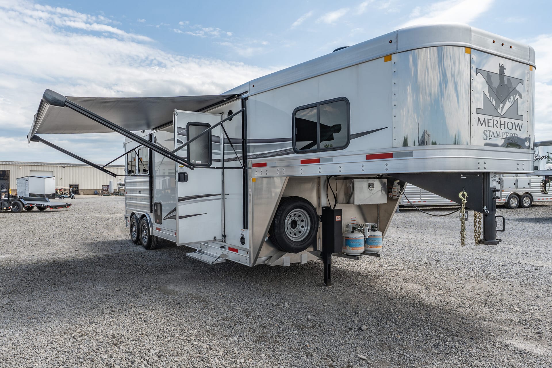 Silver Merhow Stampede horse trailer with extended awning, parked on a gravel lot under a blue sky with clouds