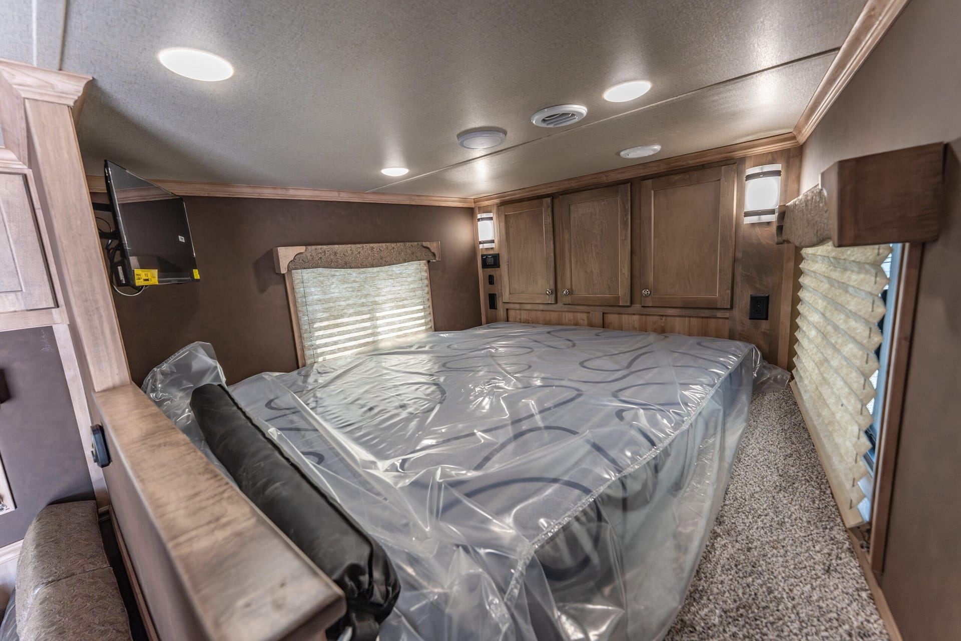 Interior view of an RV bedroom with a queen bed covered in plastic, wooden cabinets, and a mounted TV on the left wall.