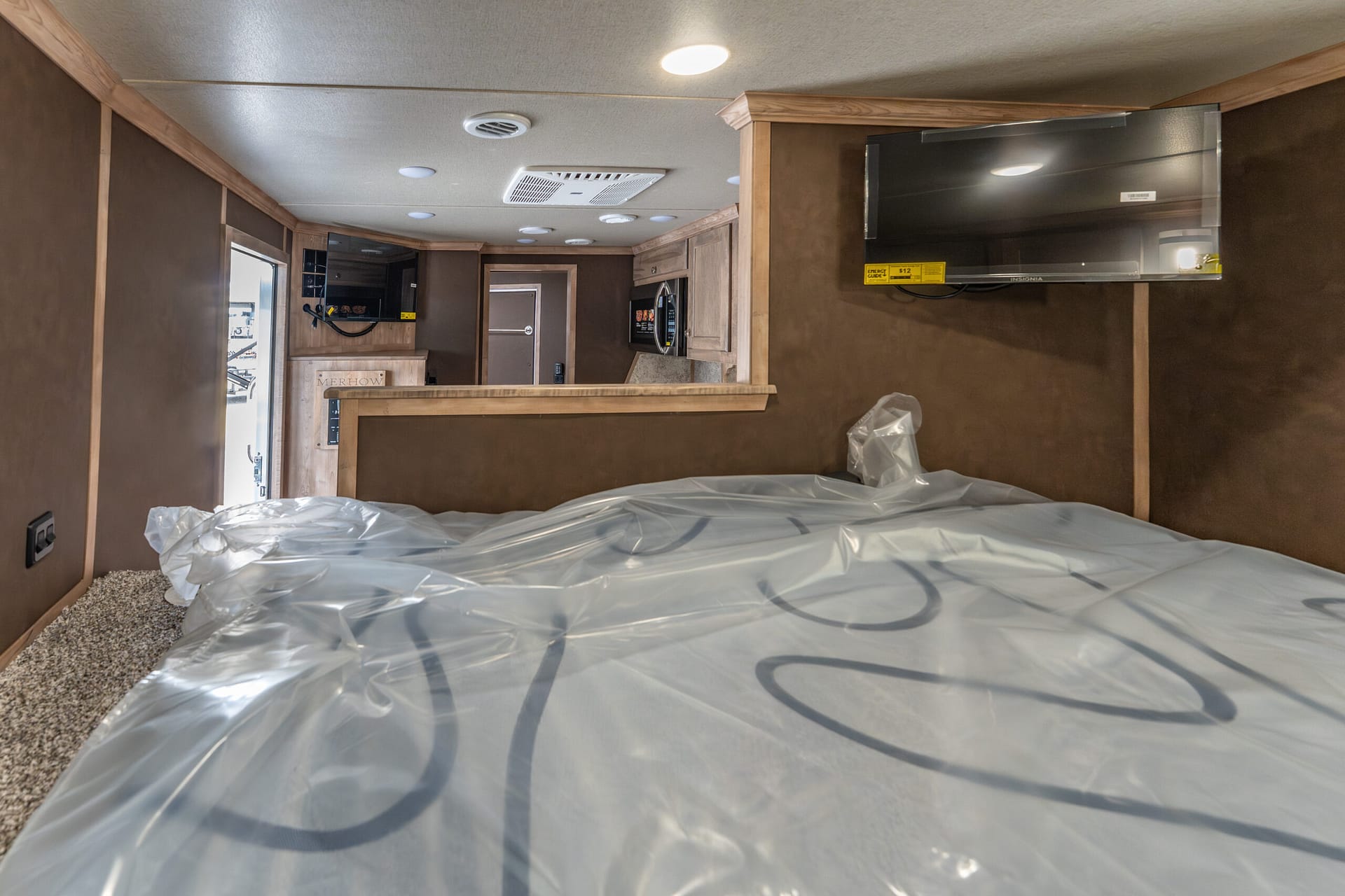 Bedroom area inside a travel trailer/RV with a bed covered in plastic, brown wood trim, and a wall-mounted TV.