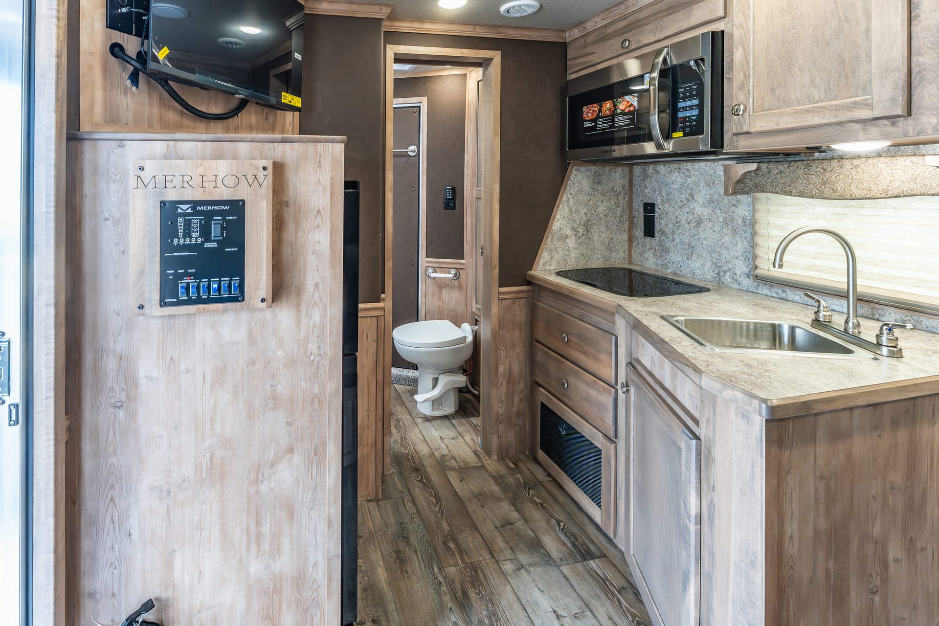 Wood-paneled RV kitchen with microwave, countertop stove, and sink; doorway leading to small bathroom with toilet in back.