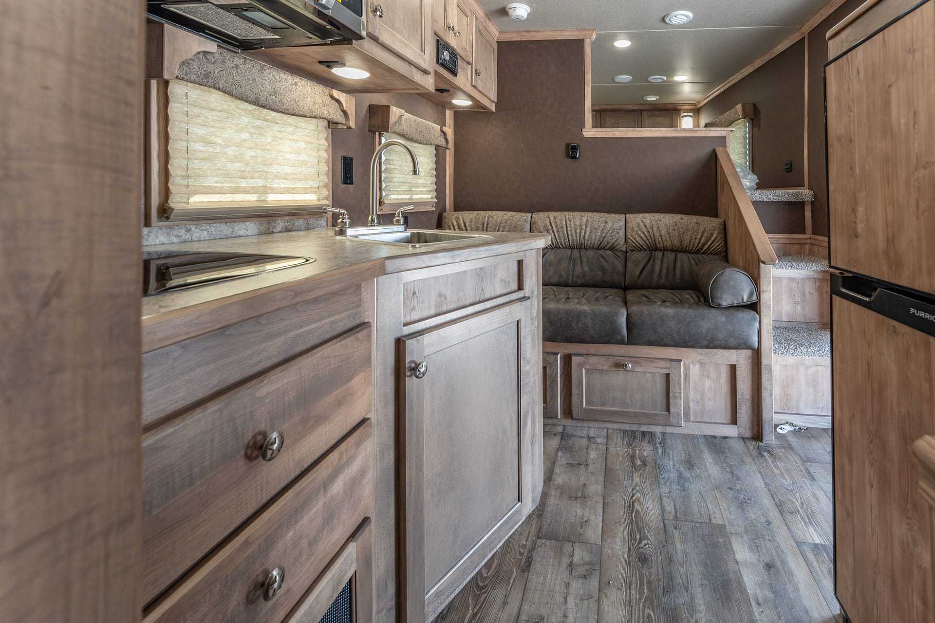 Interior of a wood-toned RV kitchen and living area with a double sink, curved faucet, and brown leather sectional sofa