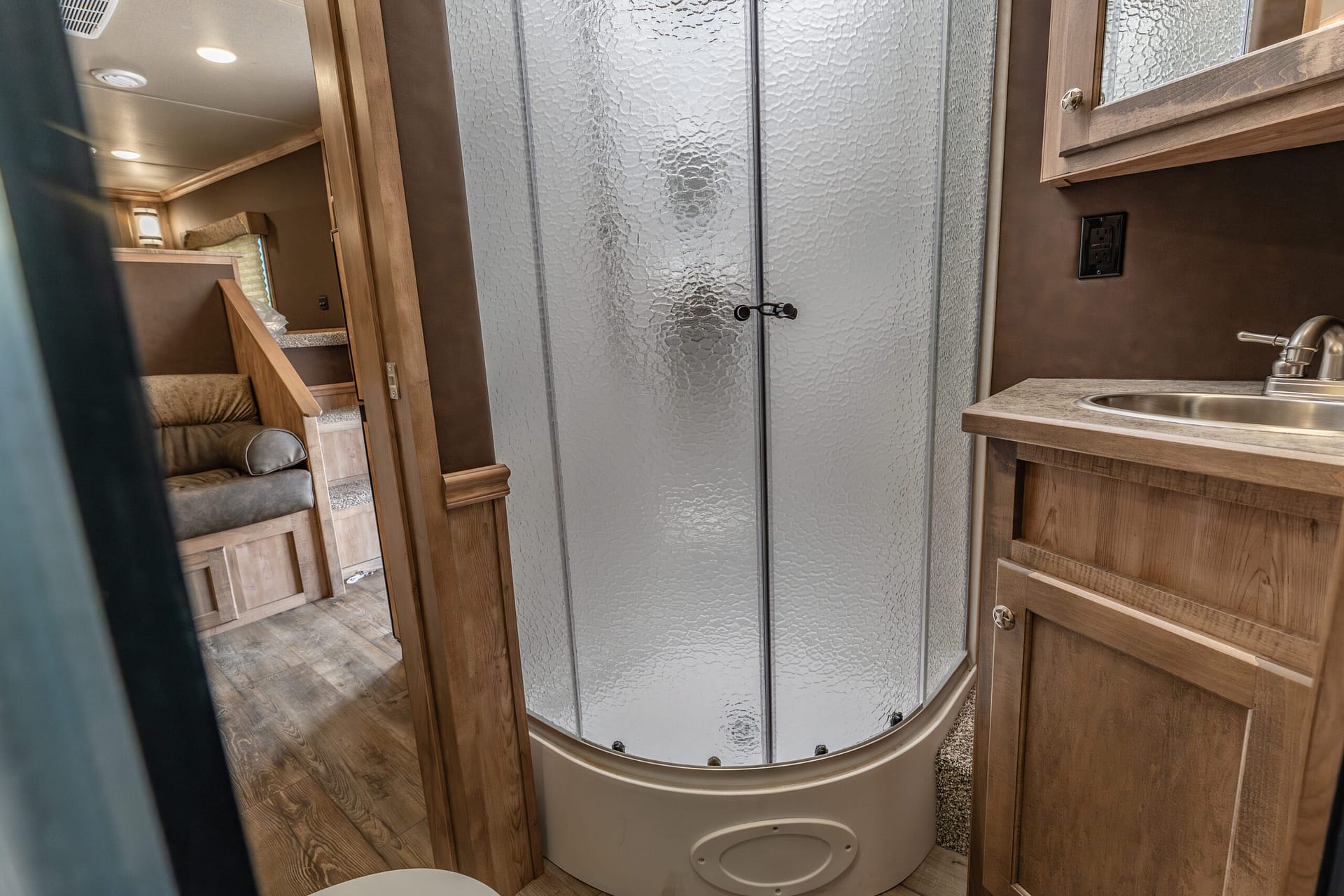 Compact RV bathroom with curved frosted-glass shower doors, wood cabinetry, and a sink along a beige countertop.