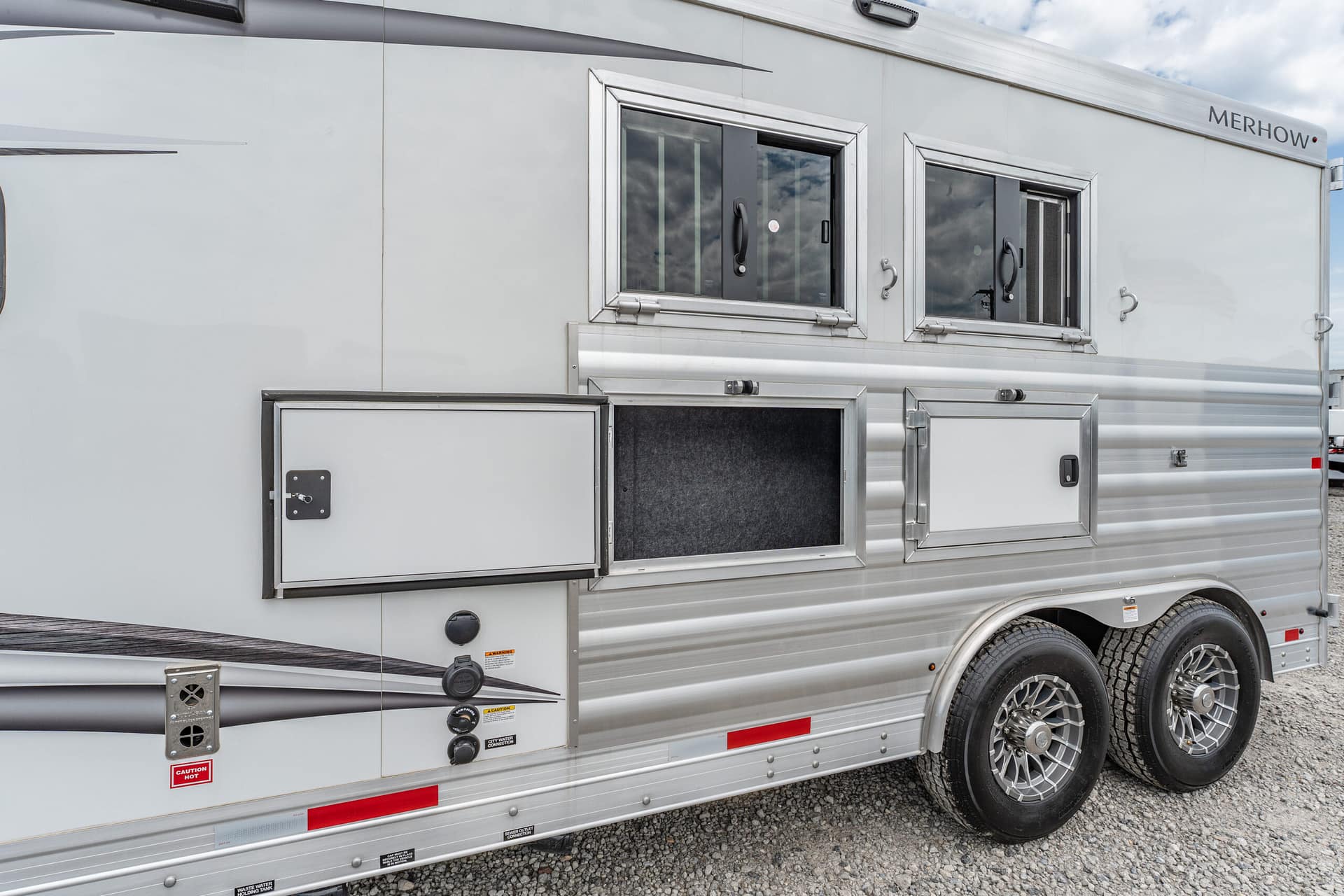 Silver horse trailer side with two framed windows, external storage compartments, and chrome trim. Dual rear wheels on gravel.