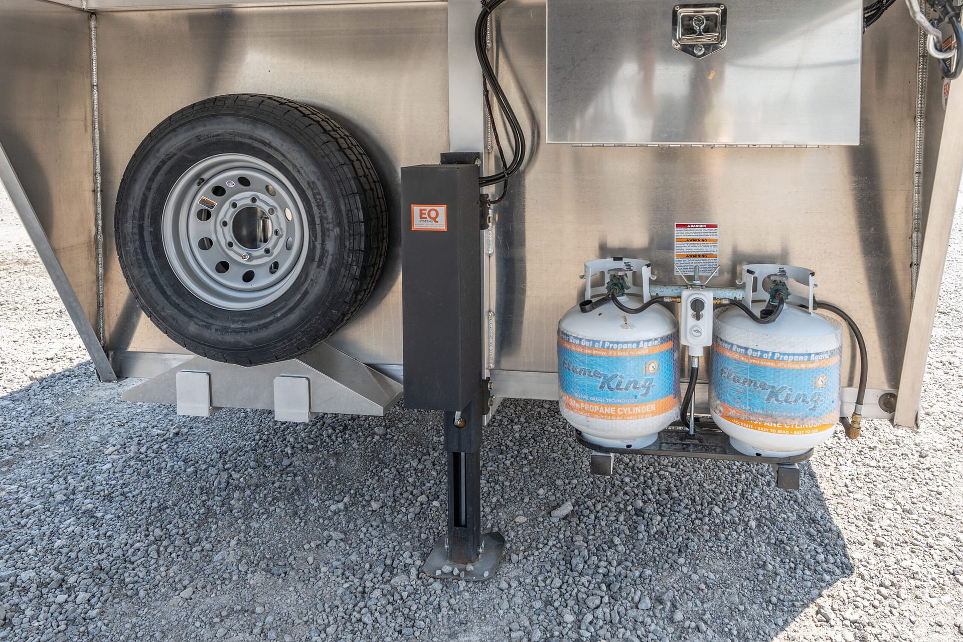 Rear side of a metal trailer with a mounted spare tire, propane cylinders, and gas connections on gravel ground.