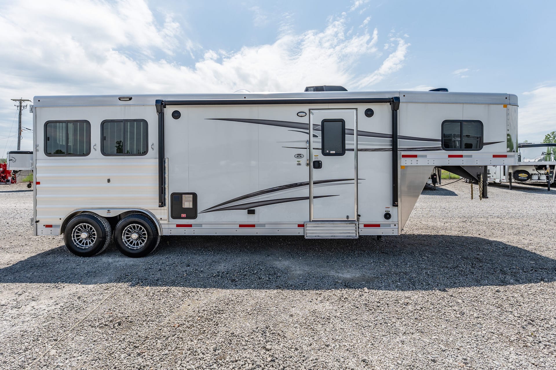 Long silver travel trailer parked on a gravel lot under a blue sky with scattered clouds, showing windows and entry door on the side