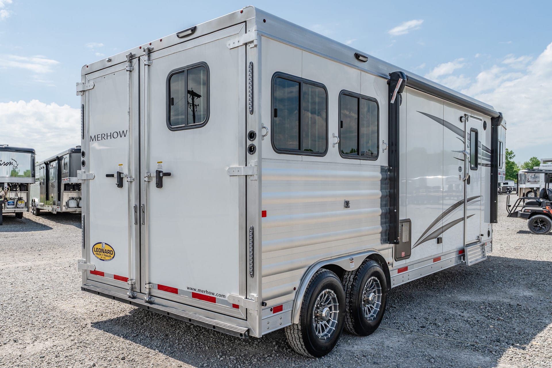 Silver Merhow horse trailer with multiple windows viewed from the back-left corner, parked on gravel lot.