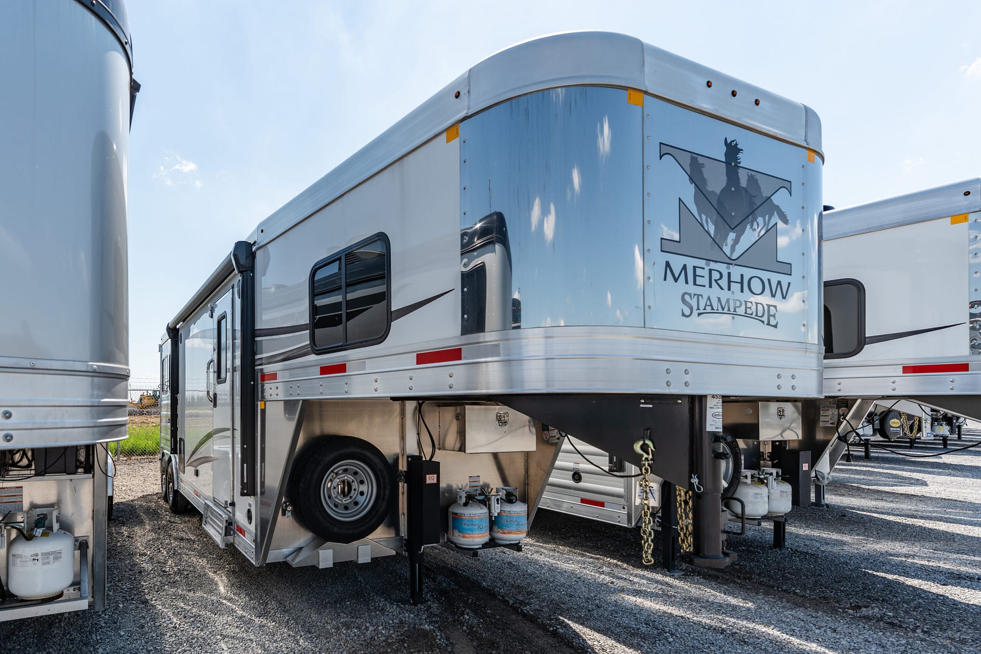 Silver Merhow Stamped horse trailer with logo on the rear, parked in a lot beside another trailer, under a blue sky.