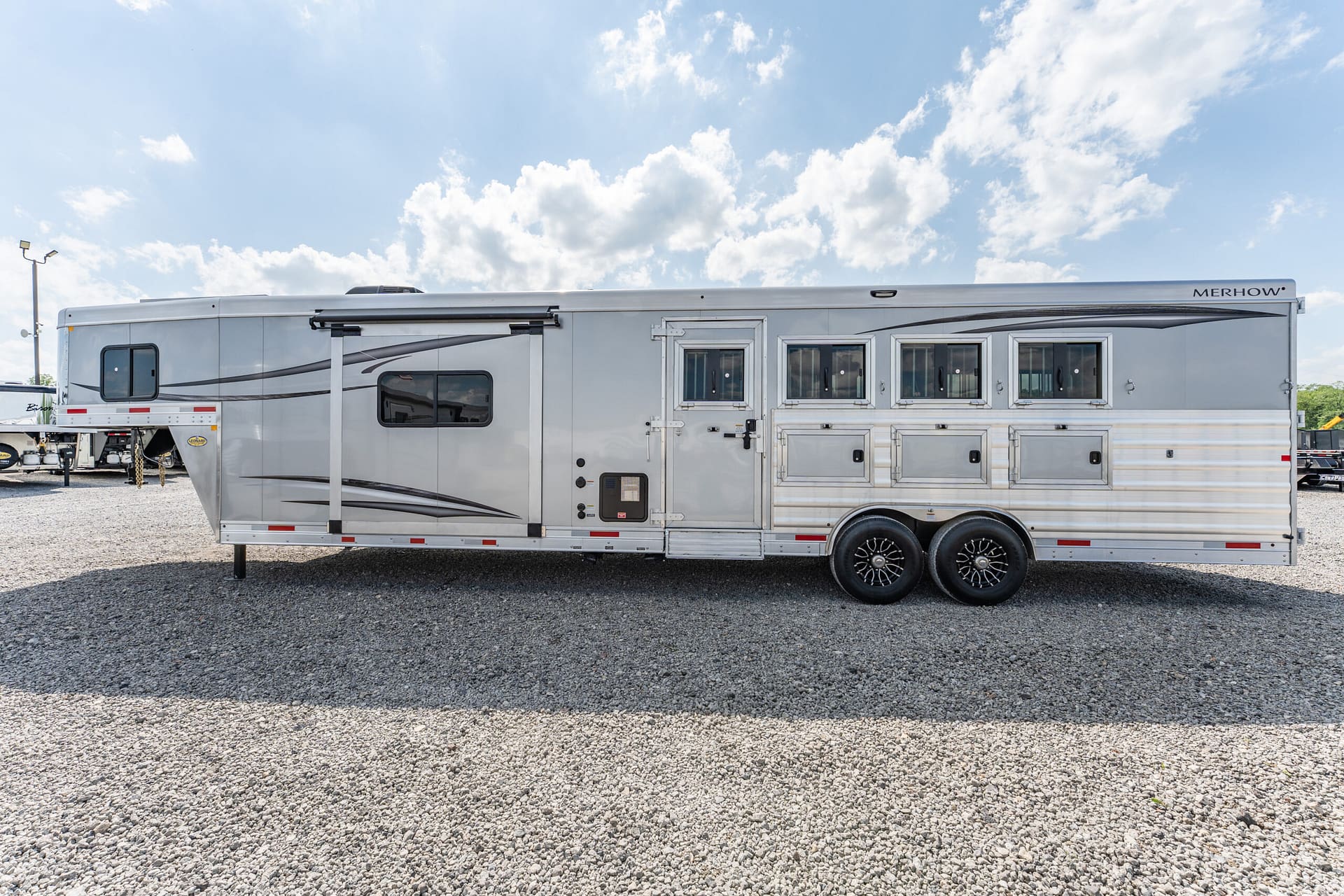 Long silver horse trailer with multiple windows and a side door, parked on a gravel lot under a blue, partly cloudy sky.