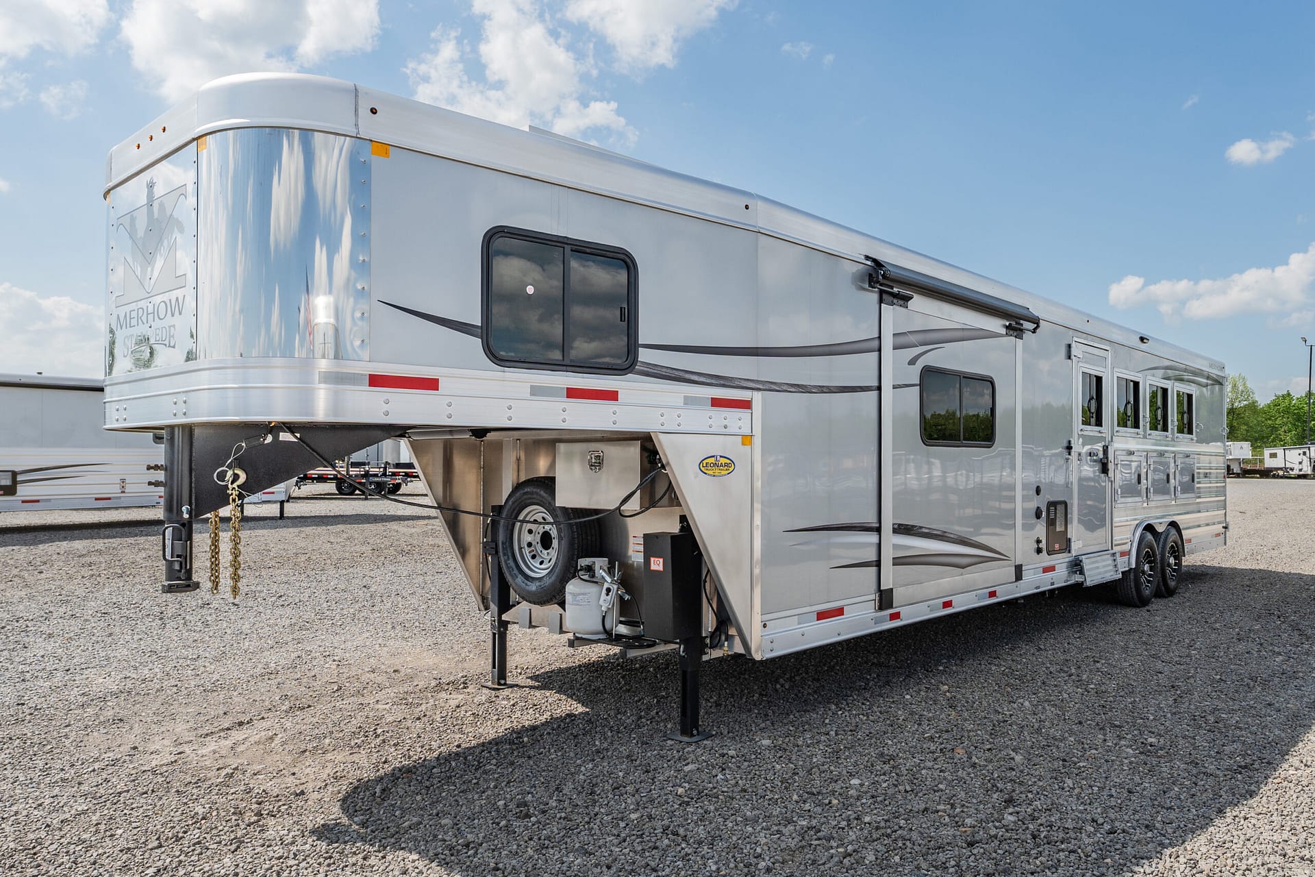 Silver long travel trailer with a gooseneck hitch parked on a gravel lot under a blue sky, showing a side window and exterior design striping.