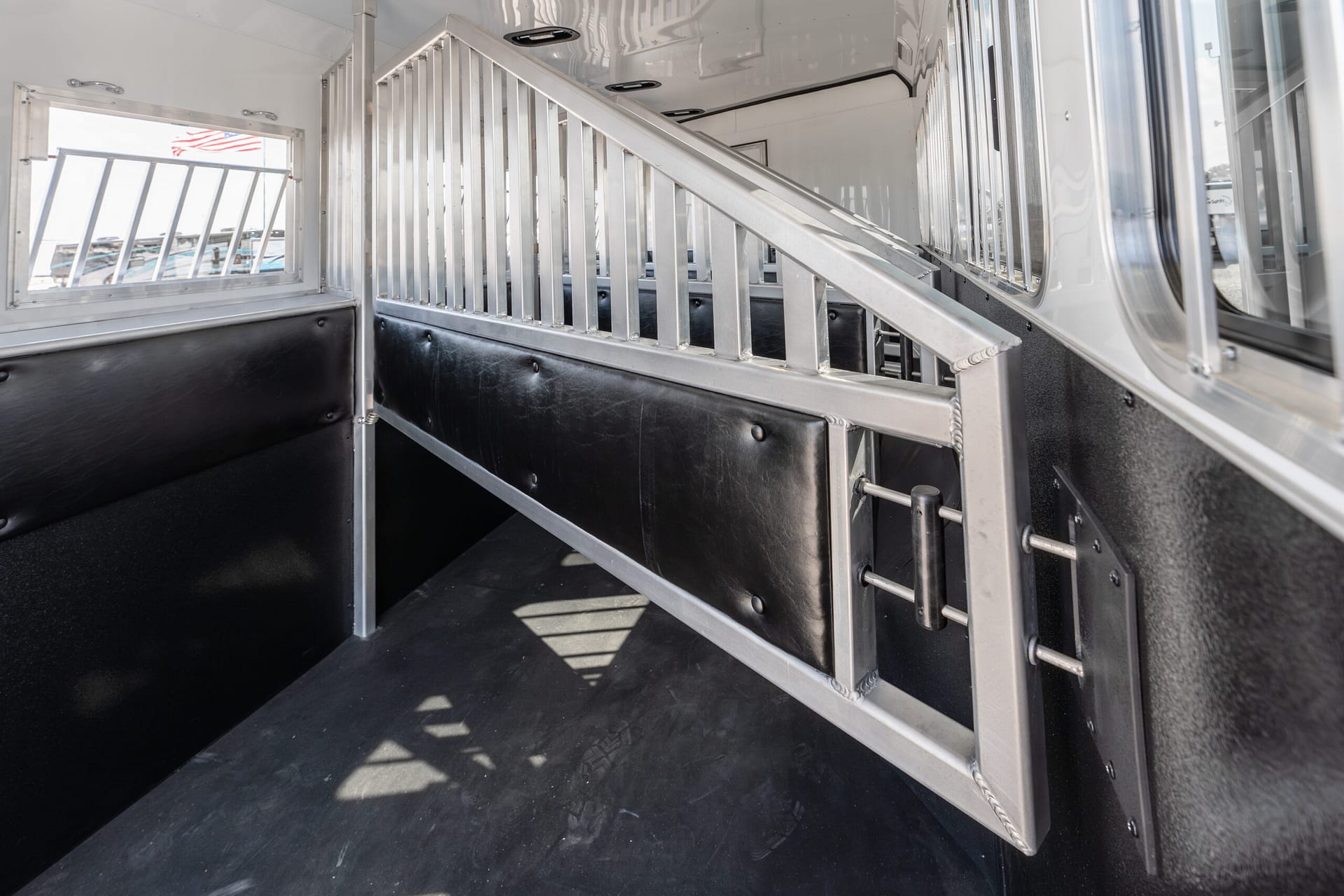 Interior of a metal horse trailer with a diagonal divider, aluminum rails, black padded walls, and a window showing an American flag outside.