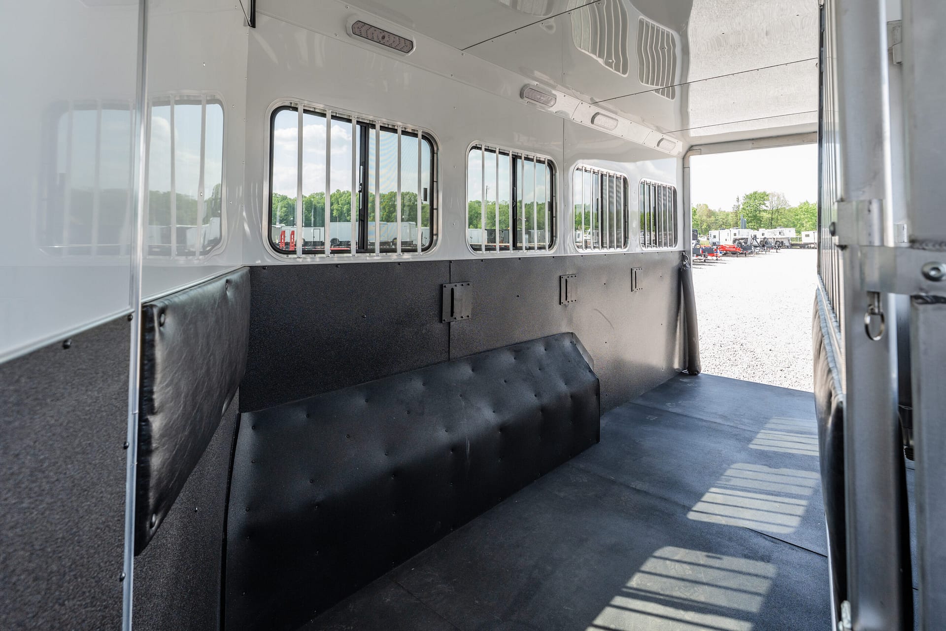 Interior of a metallic trailer with padded black walls and barred windows, opened to outdoor parking area with trailers beyond.