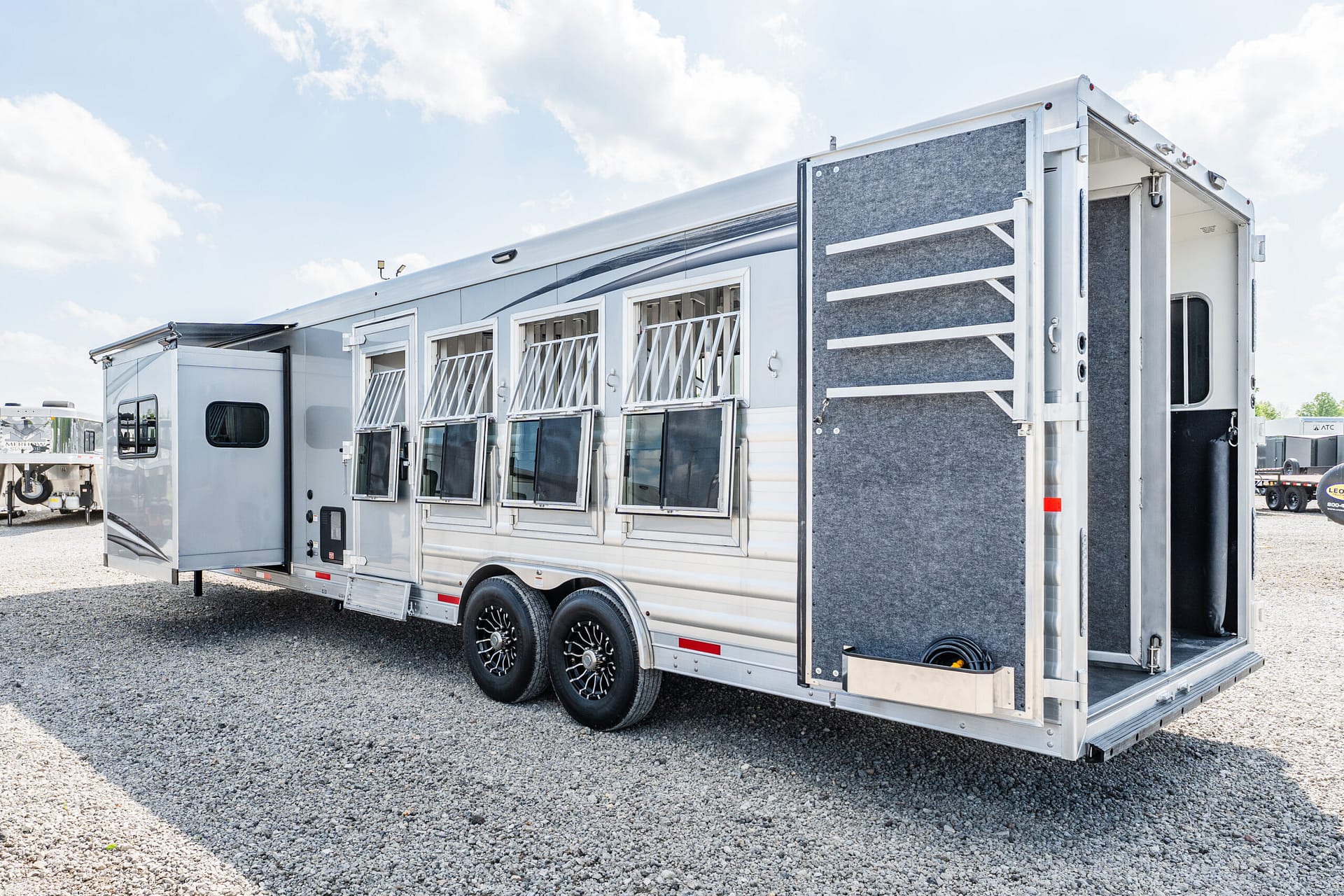 White horse trailer with side windows and an open rear ramp on a gravel lot under a blue sky