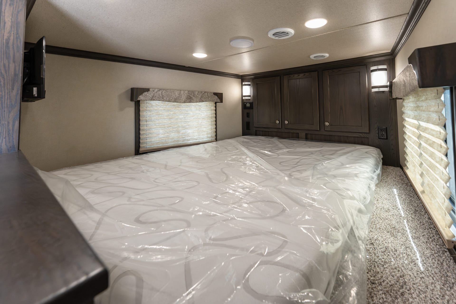 Interior of a compact RV bedroom with a large bed in the foreground, dark wood cabinets along the back wall, and a window with beige blinds to the left. Ceiling lights illuminate the space.