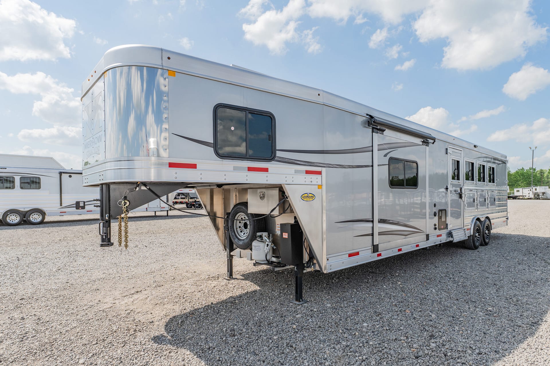 Silver gooseneck travel trailer on a gravel lot, with a side window and spare wheel visible under a blue sky.