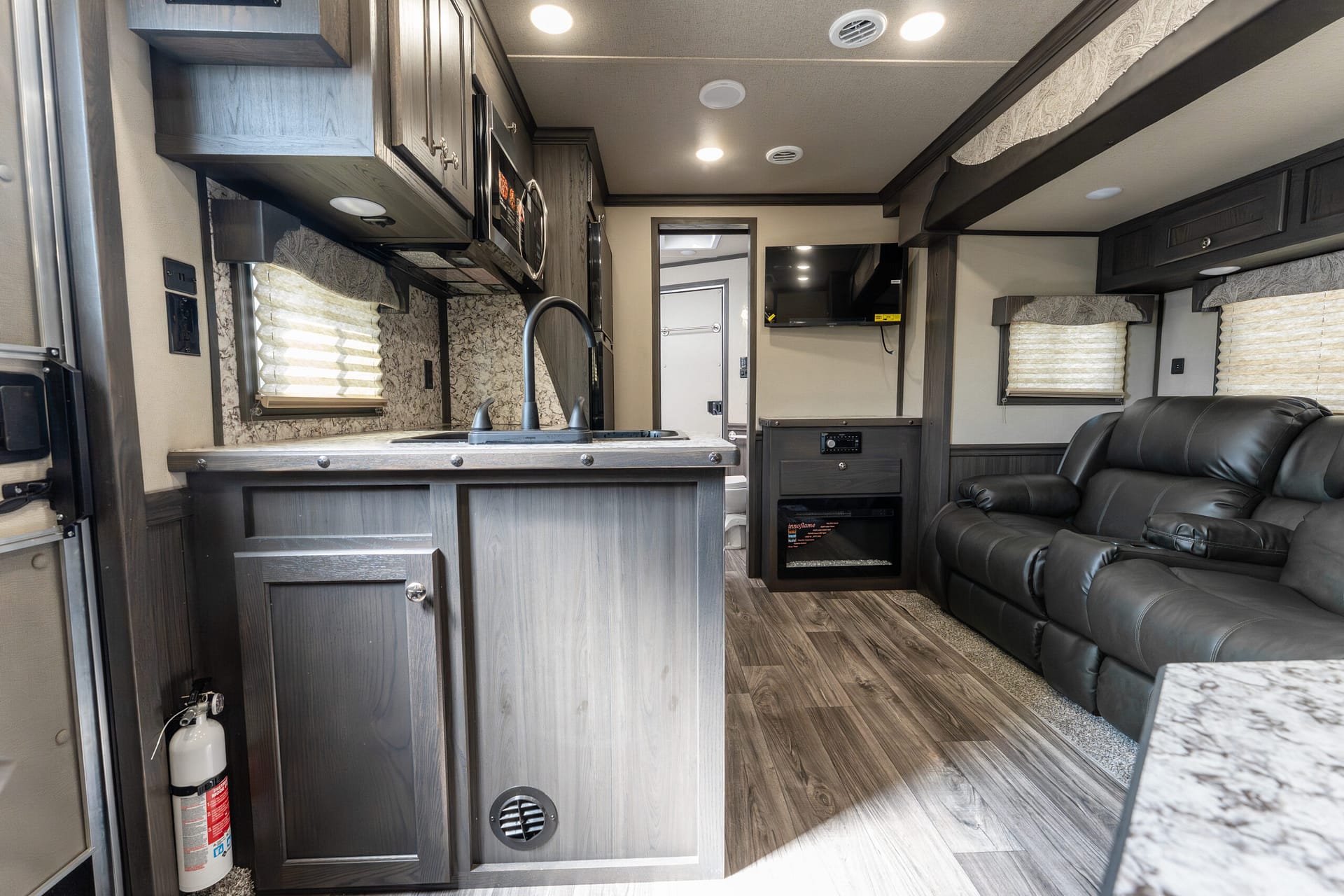 Interior of a modern RV kitchen and living area with gray wood cabinets, marble-like countertop, black sink faucet, and a leather reclining sofa nearby.