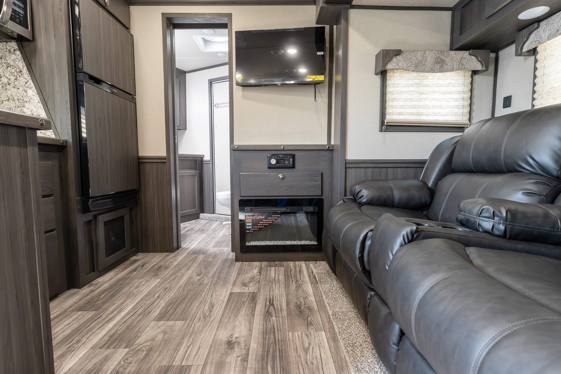 Interior of a modern RV living area with dark wood cabinets, a wall-mounted TV, and a leather sectional sofa across from the screen.