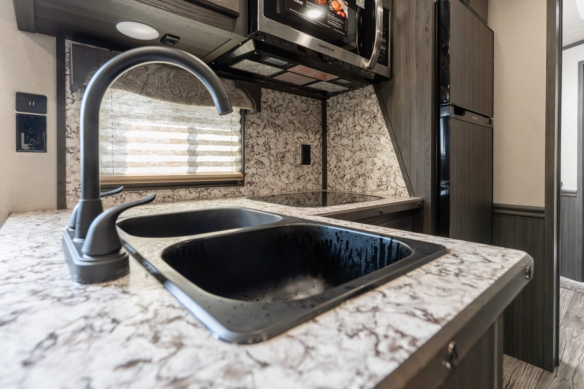 RV kitchen counter with black sink, curved faucet, and marble-like countertop beneath dark cabinets and a window shade.