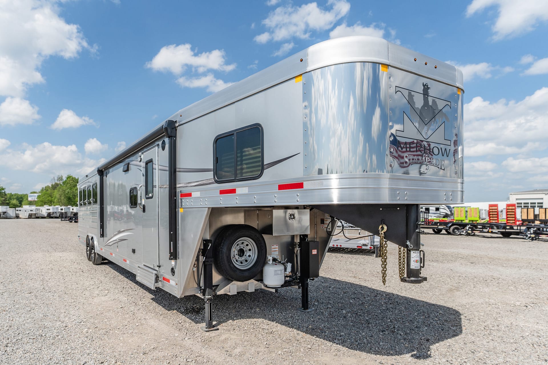 Silver aluminum gooseneck horse trailer parked on a gravel lot under a blue sky, stabilizing legs deployed and logo visible on the rear corner.