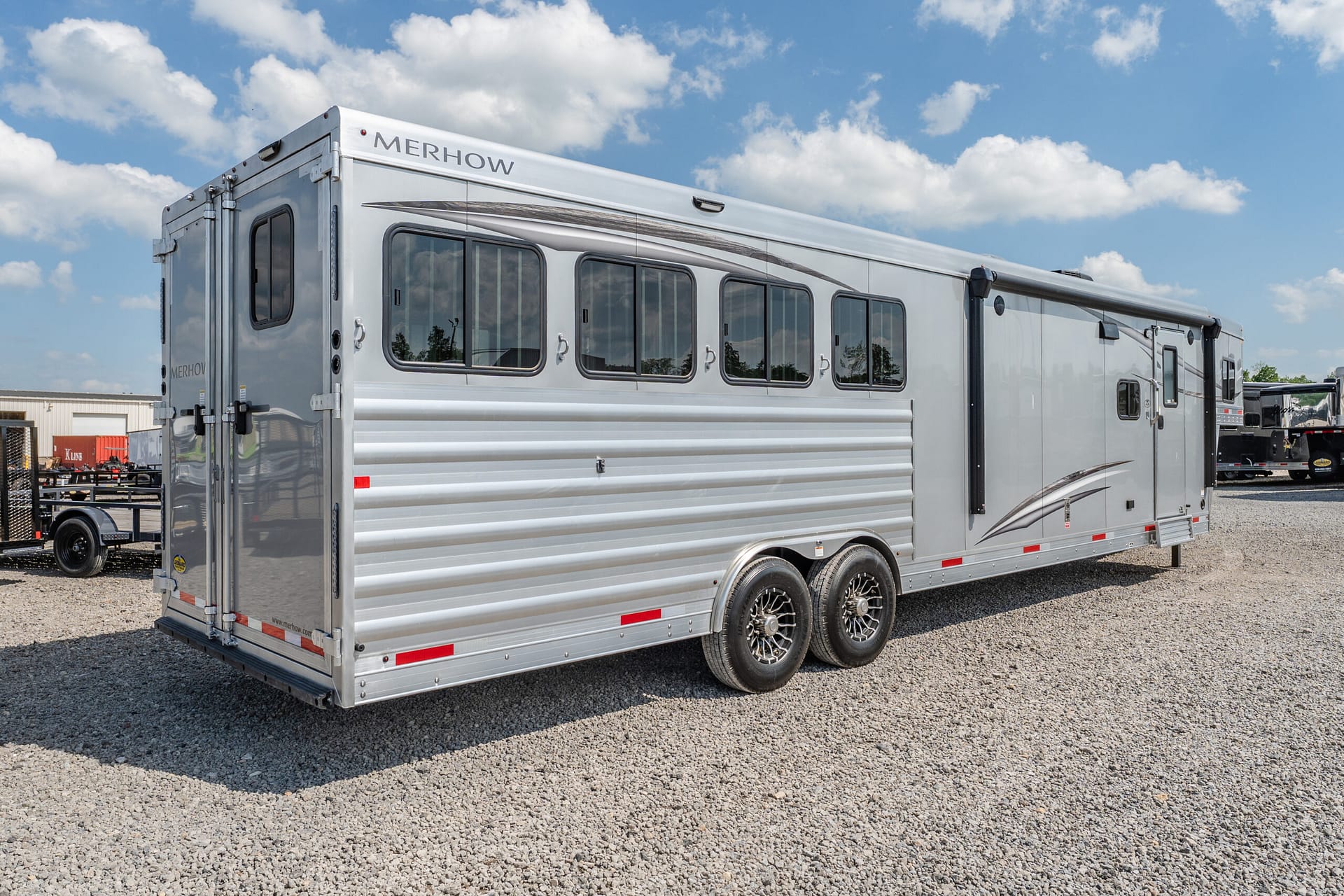 Silver Merhow horse trailer with multiple windows, parked on a gravel lot under a blue sky.