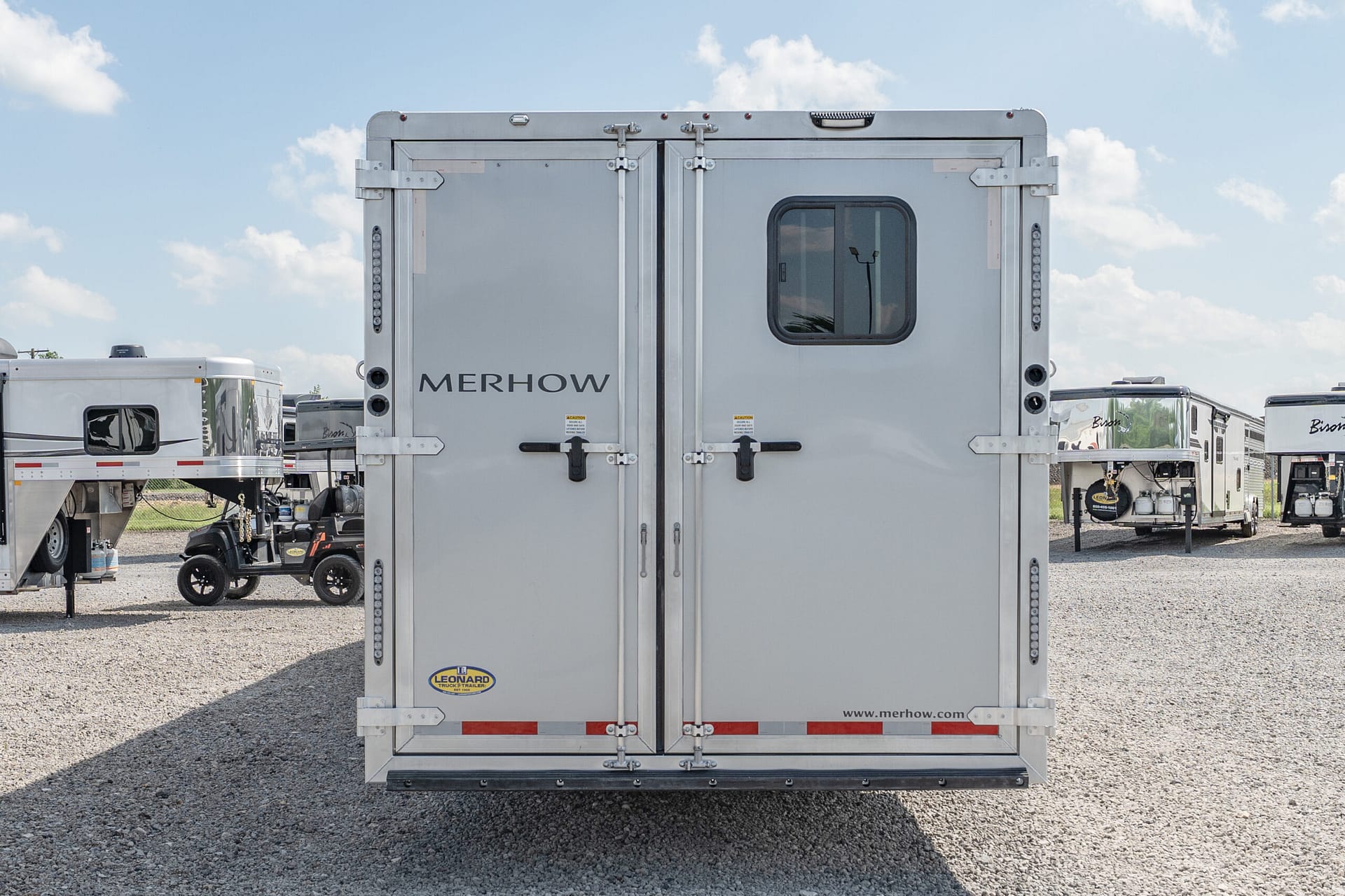 Rear view of a silver Merhow horse trailer with double doors and a small window, parked among other trailers on a gravel lot under a blue sky.