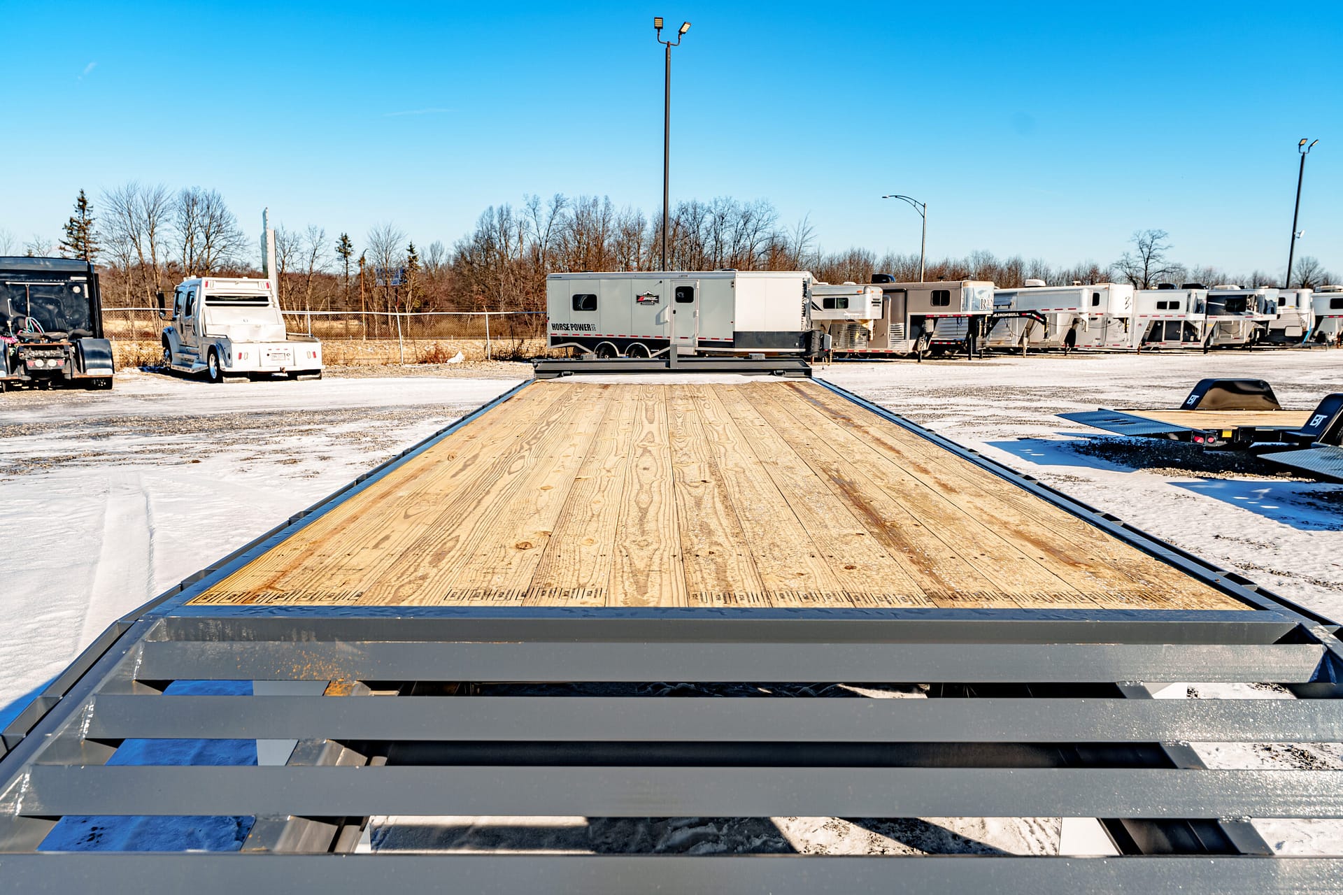 Wide view of a row of parked semi-trailers in a snow-dusted lot under a clear blue sky, seen from a wooden flatbed in the foreground.
