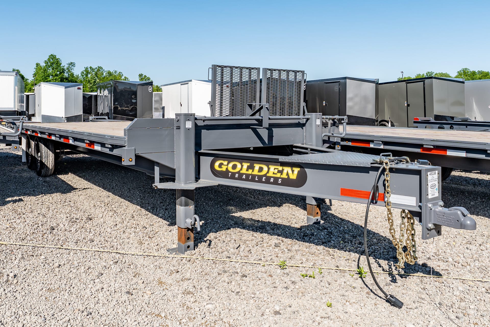 Grey flatbed trailer with a metal ramp and chain ties in a gravel lot, branded 'Golden Trailers', with storage trailers in the background.