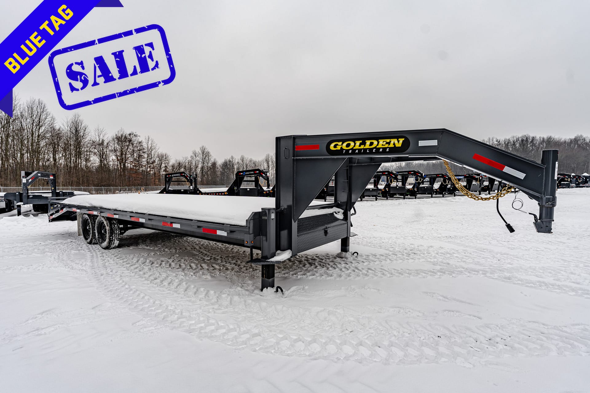 Blue sale tag diagonally across snowy lot with stacked trailers labeled Golden Trailers in background.