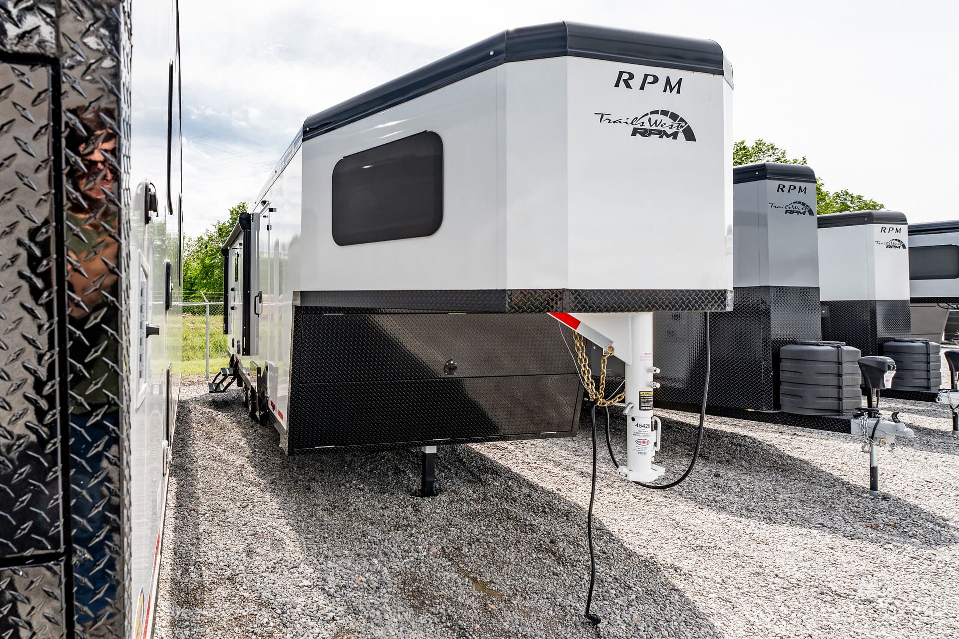 Row of white RP M Trails West travel trailers with black diamond plate skirts parked on a gravel lot, facing a chain-link fence and trees in the background.