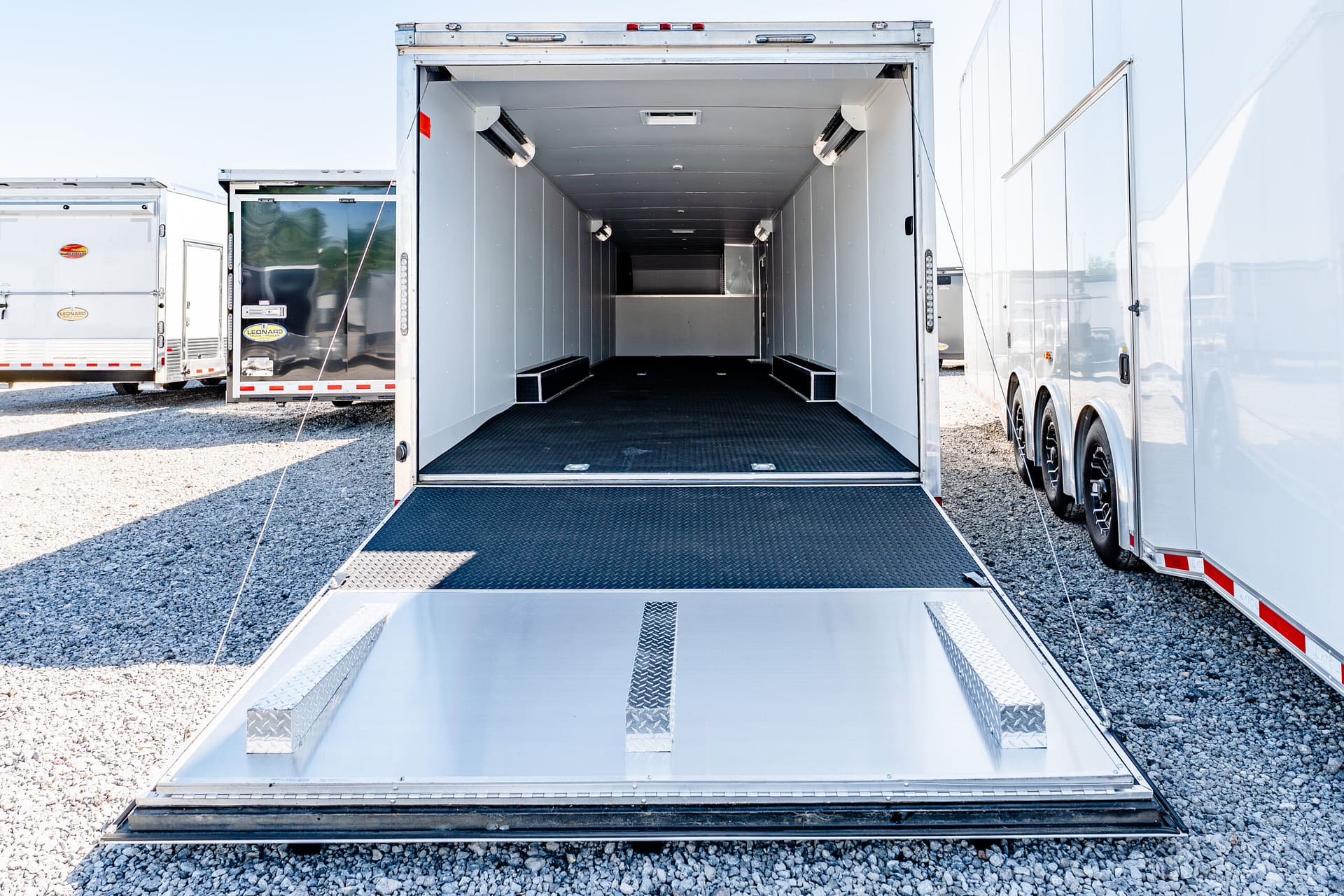 Open empty enclosed trailer with diamond-plate ramp down, parked on a gravel lot beside other trailers.