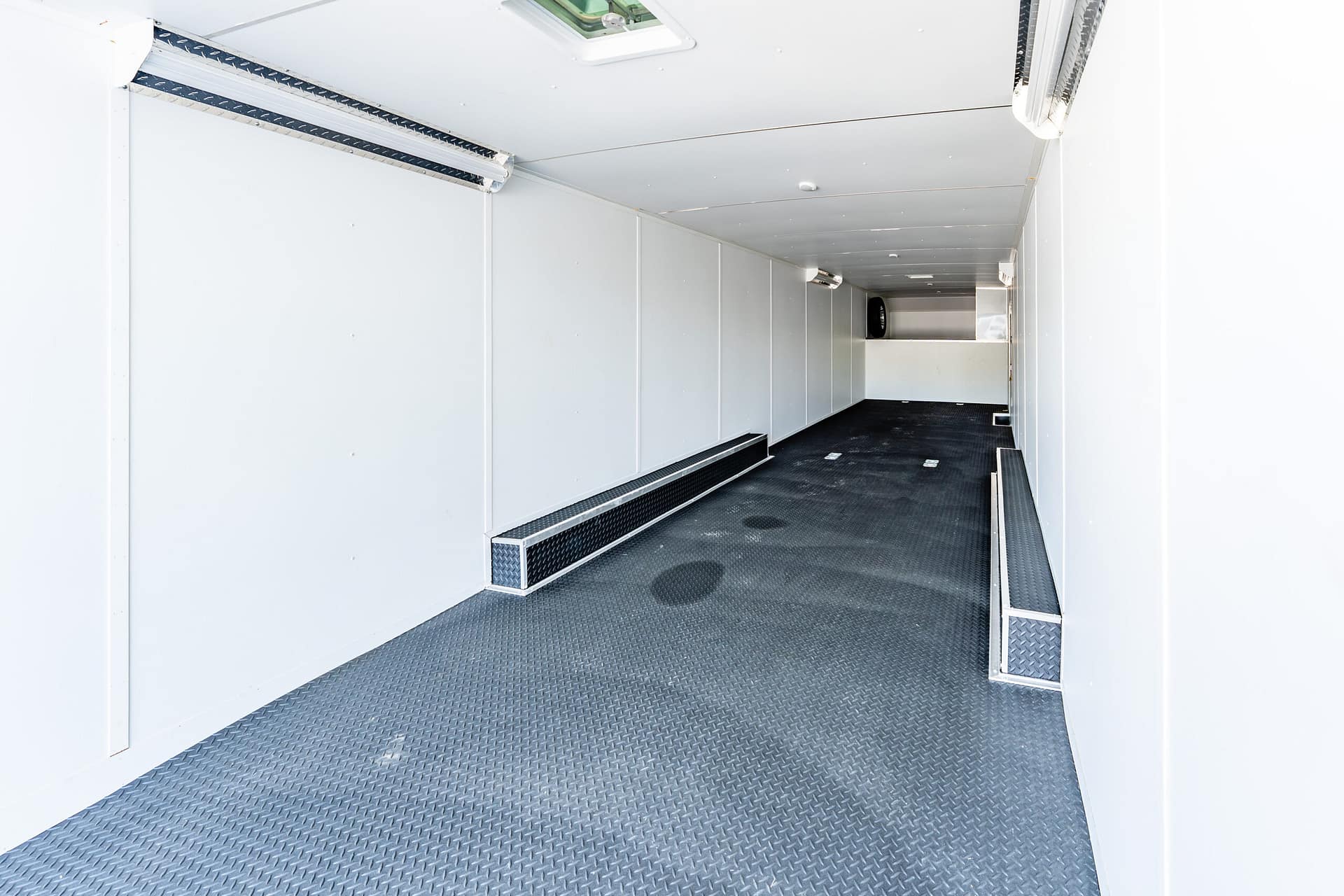 Inside a white cargo trailer with a diamond-plate floor, overhead lights, and a spare tire visible at the far end.