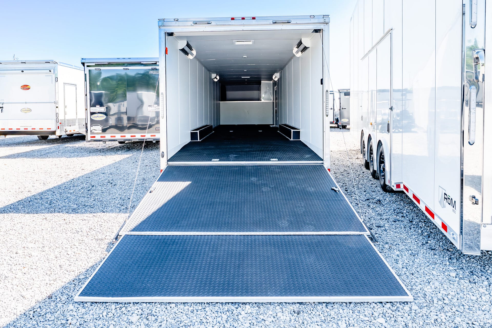 Open cargo trailer with a lowered metal ramp leading into an empty interior, parked among other trailers in a gravel lot.