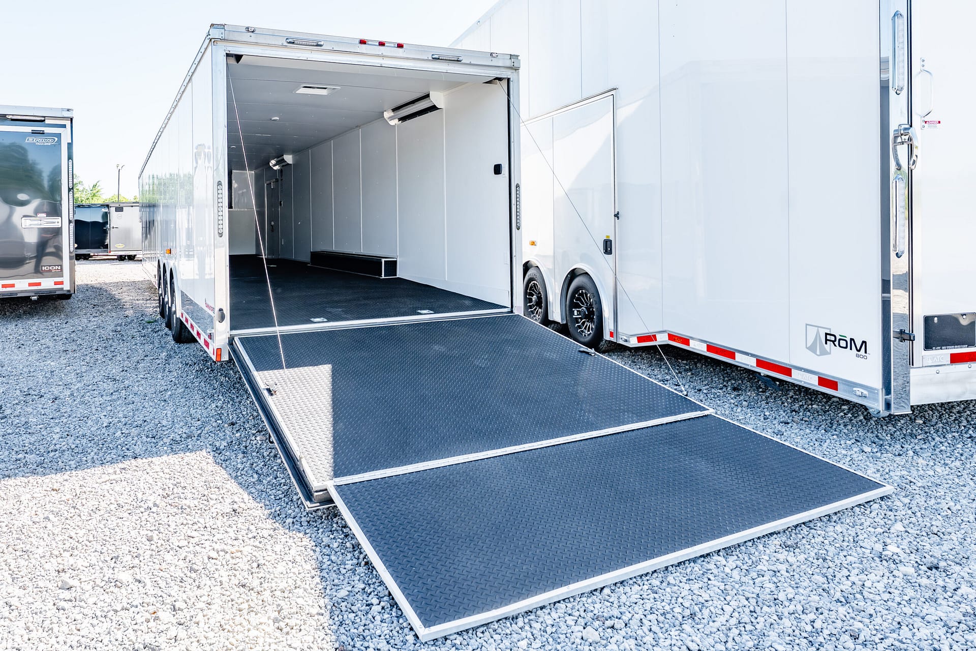 White semi-trailer with its back doors open and a black diamond-plate loading ramp extended onto a gravel lot.