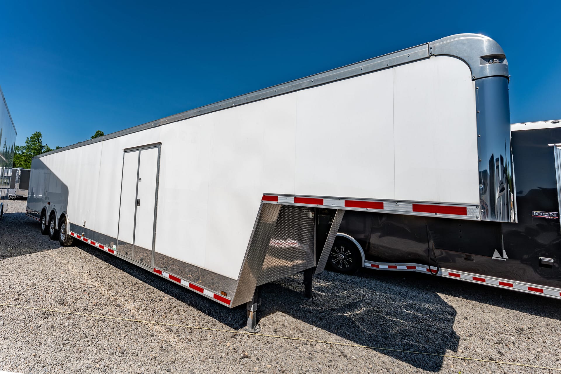 Long white semi-trailer with chrome accents and diamond-plate ramp area, parked on a gravel lot under a clear blue sky
