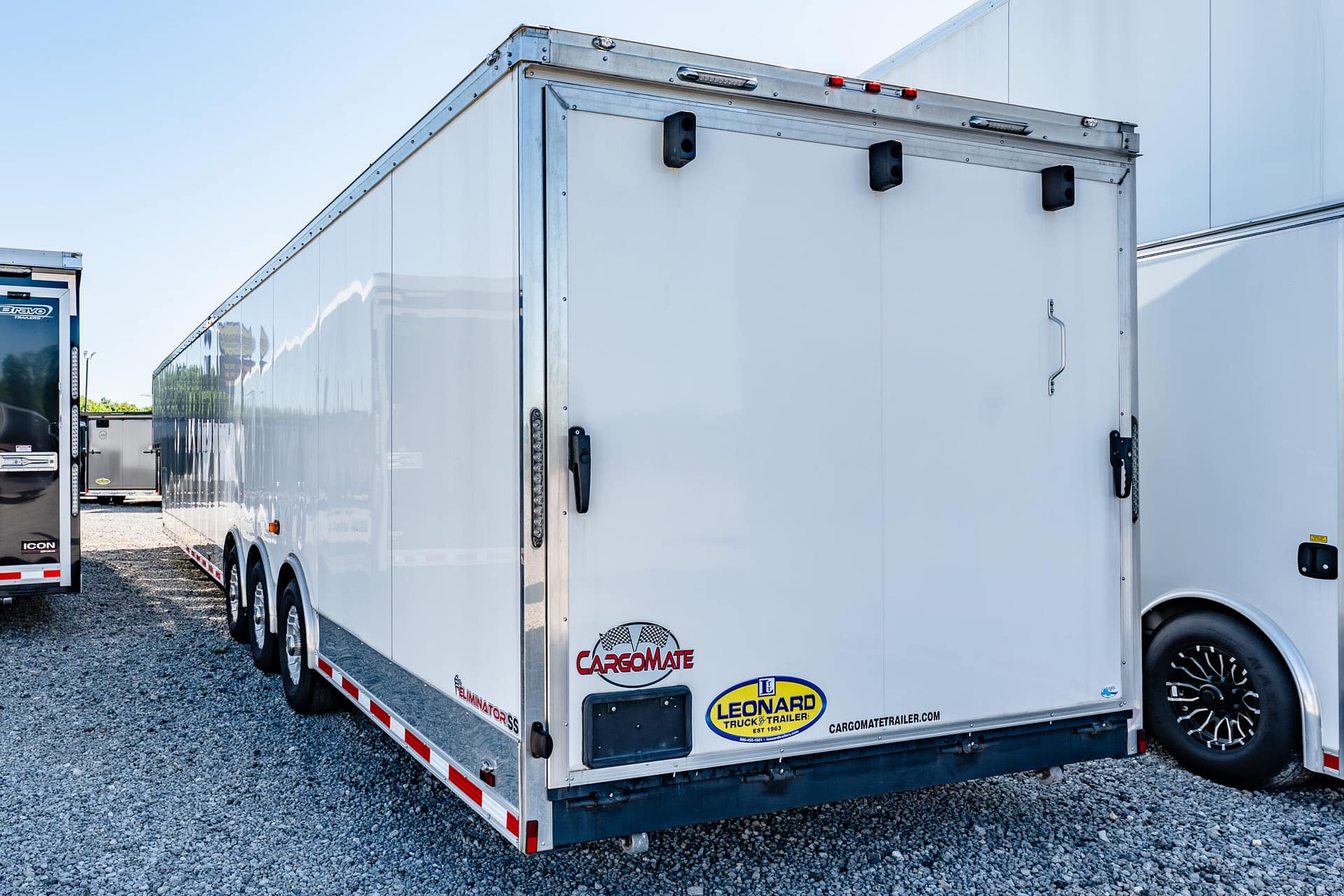 Large white cargo trailer with branding decals (Cargomate, Leonard) parked on a gravel lot, another trailer visible in the background on the left cap of the image.