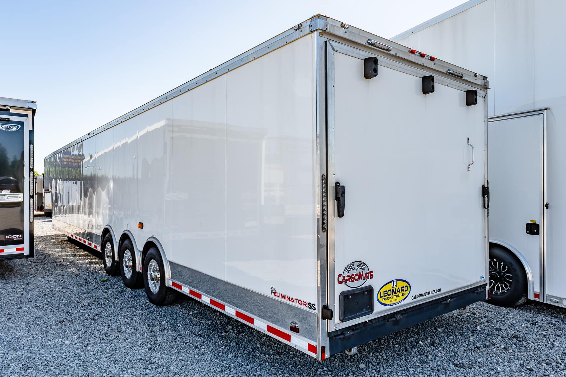 Side view of a long white cargo trailer with chrome trim parked on gravel, with another trailer visible at the left edge and various decals near the bottom.