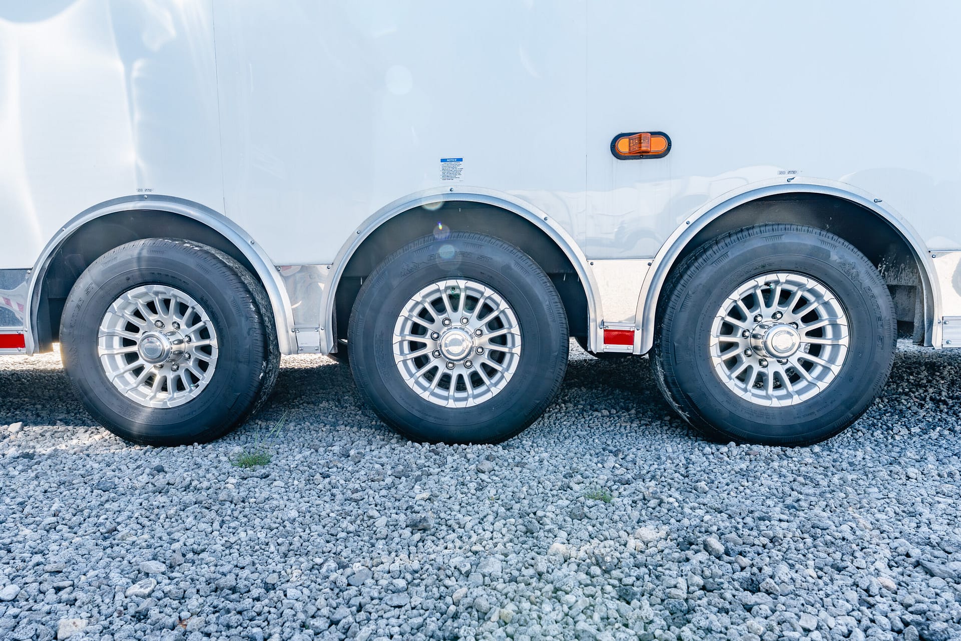 Triple trailer wheels with silver alloy rims mounted on a white trailer, parked on gravel.