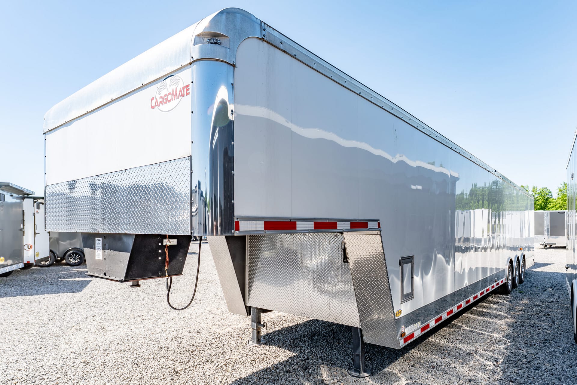 Shiny silver cargo trailer with diamond-plate lower panels parked in a gravel lot under a clear blue sky, logo visible on the front corner.