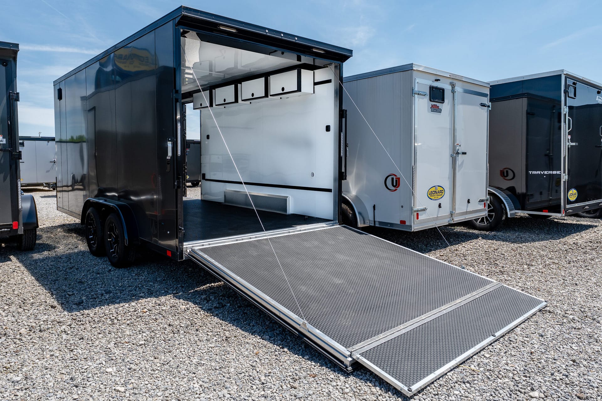Open black trailer with an extended loading ramp revealing white interior storage cabinets, parked among other trailers on gravel under a blue sky.