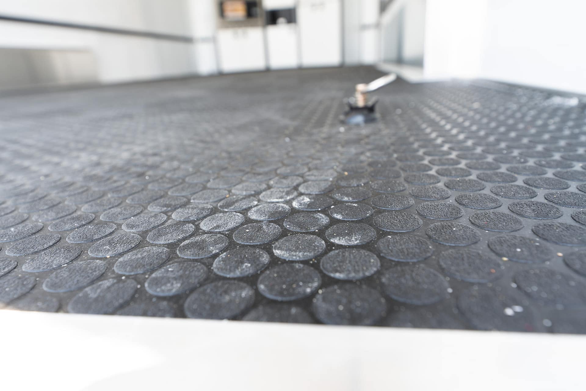Close-up of a textured black non-slip floor with circular studs, foreground shows a small metal bolt, background blurred indoors.