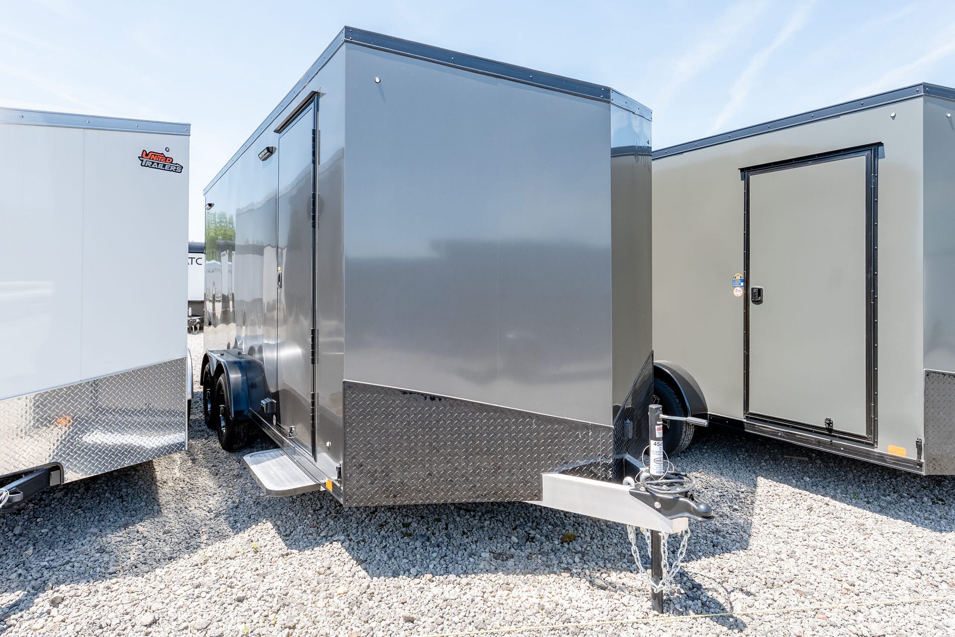Grey enclosed cargo trailers lined up on a gravel lot under a blue sky, with diamond-plate accents and a visible hitch in the foreground.