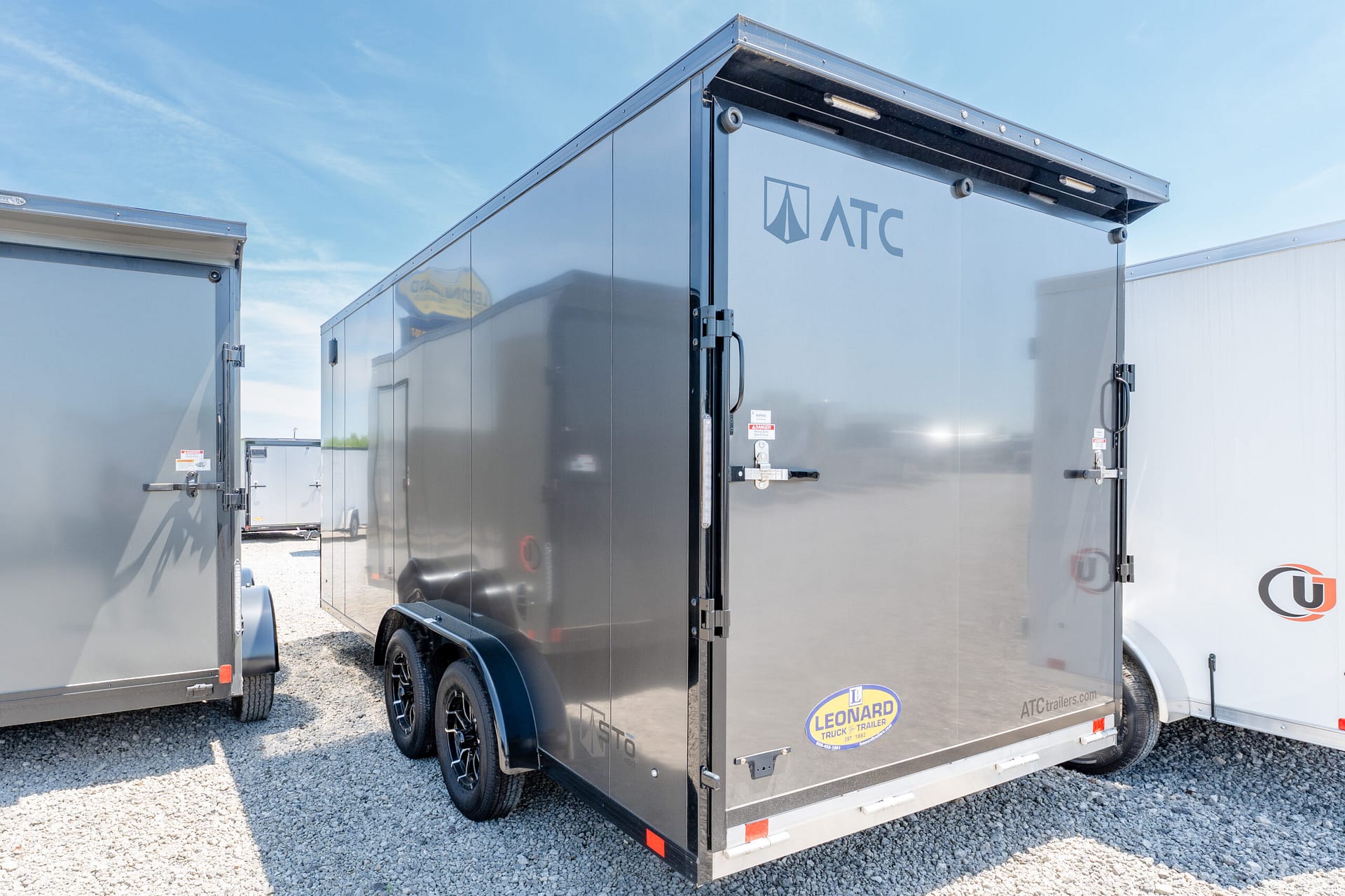 Rear corner of a shiny gray cargo trailer with ATC logo, parked among other trailers on a gravel lot.