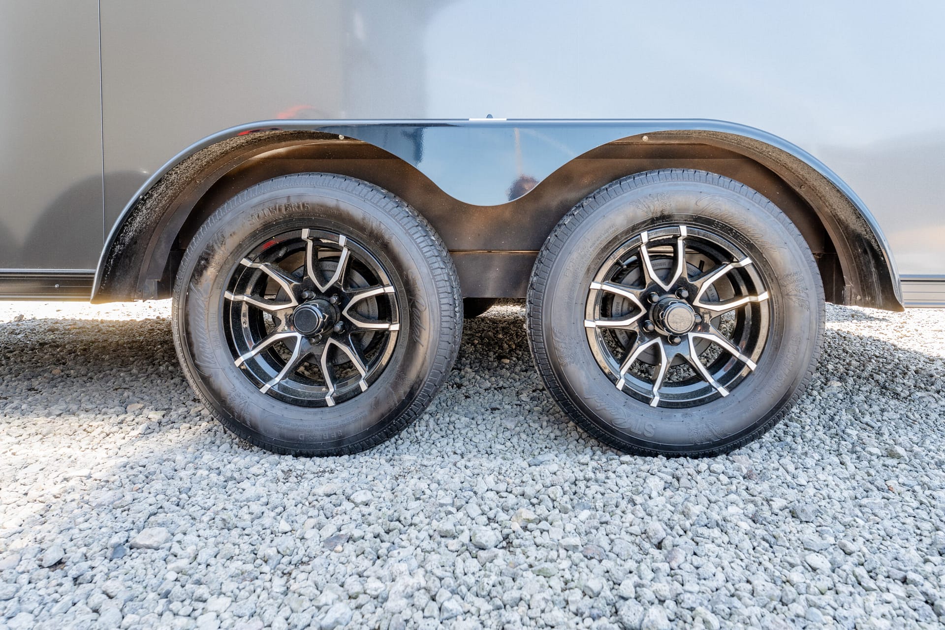 Dual-axle trailer wheel area with black alloy rims, tires, and a curved fender against a gray trailer wall on a gravel surface.
